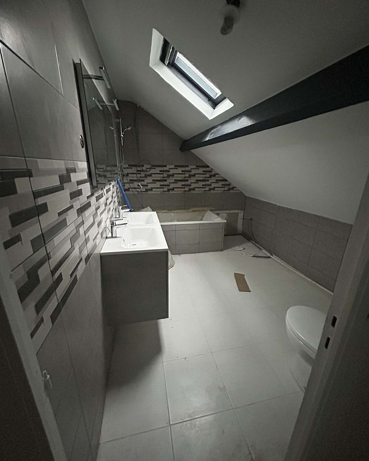 Après travaux : même pièce sous combles en carrelage clair, bandeau décoratif, plan double vasque et baignoire habillée.