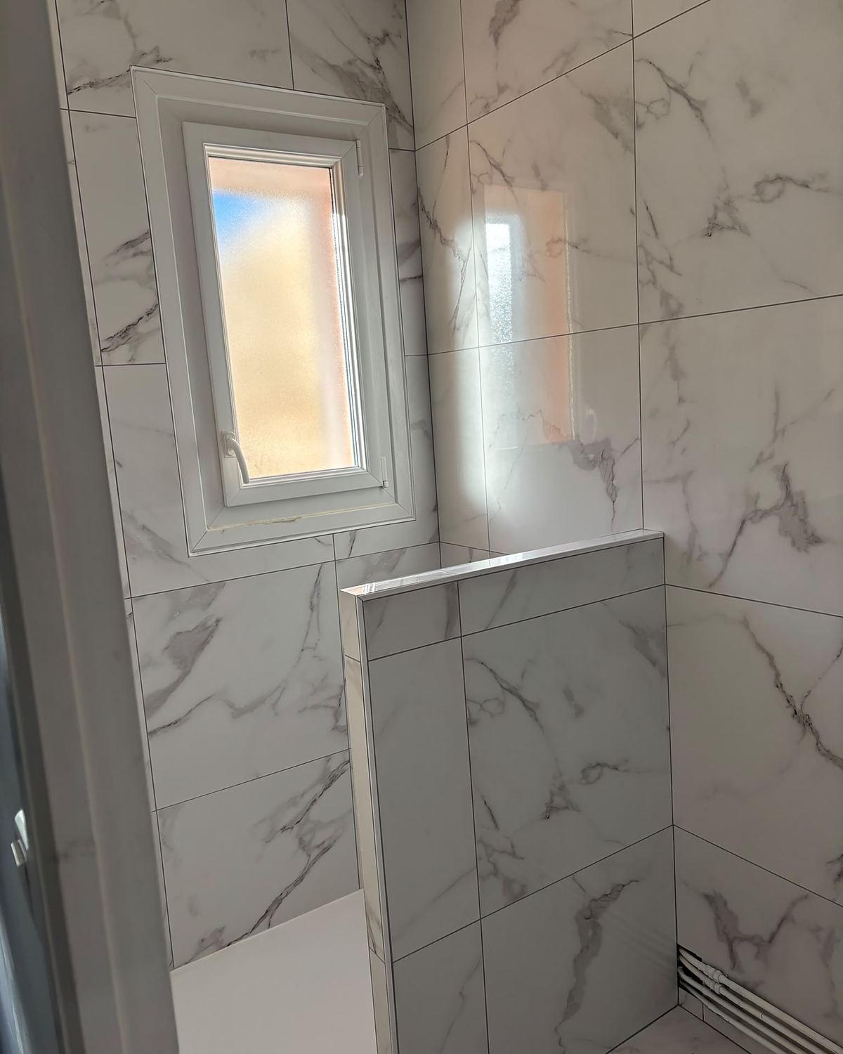 Après travaux : même salle d'eau entièrement en grès cérame effet marbre blanc, avec un muret de douche et une évacuation d'eau en longueur.