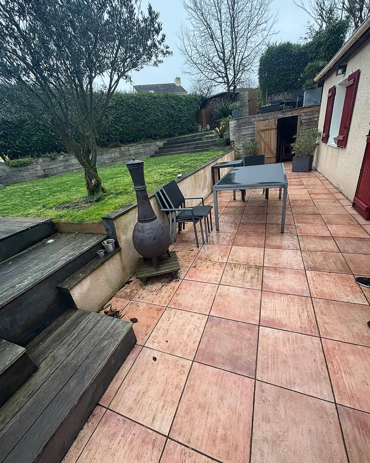 Avant travaux : terrasse en carrelage terre cuite le long de la maison, avec table de jardin et marches en bois.