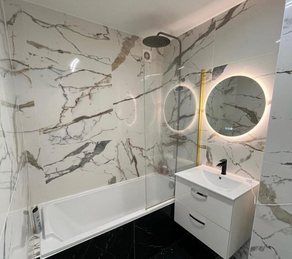Salle de bain carrelée en grand format effet marbre, sol en marbre noir, baignoire habillée et miroirs ronds rétroéclairés.