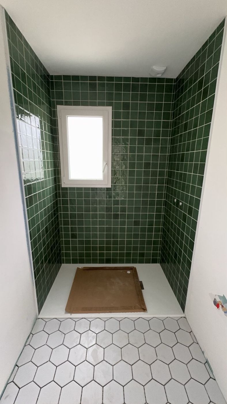 Douche carrelée en faïence vert émeraude sur trois murs, sol en carreaux hexagonaux blancs.
